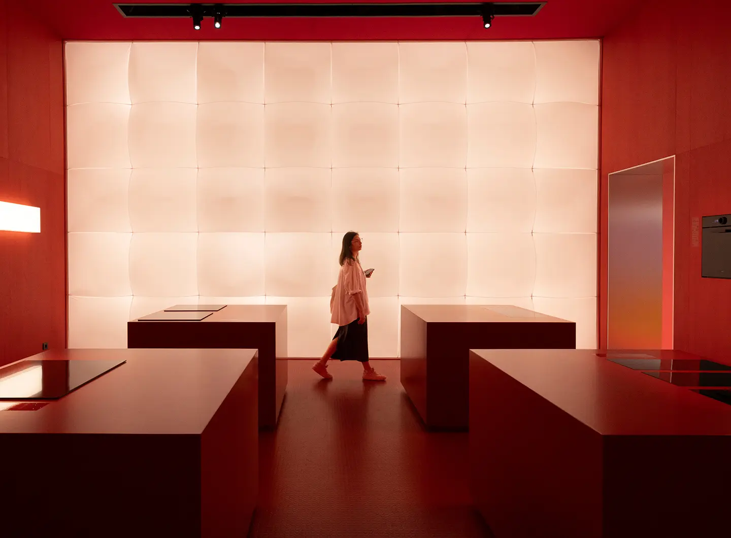Vista prospettica dello stand con al centro una ragazza e quattro banconi ai lati. Sullo sfondo un pannello di luci che illuminano la stanza i cui colori tendono al rosso