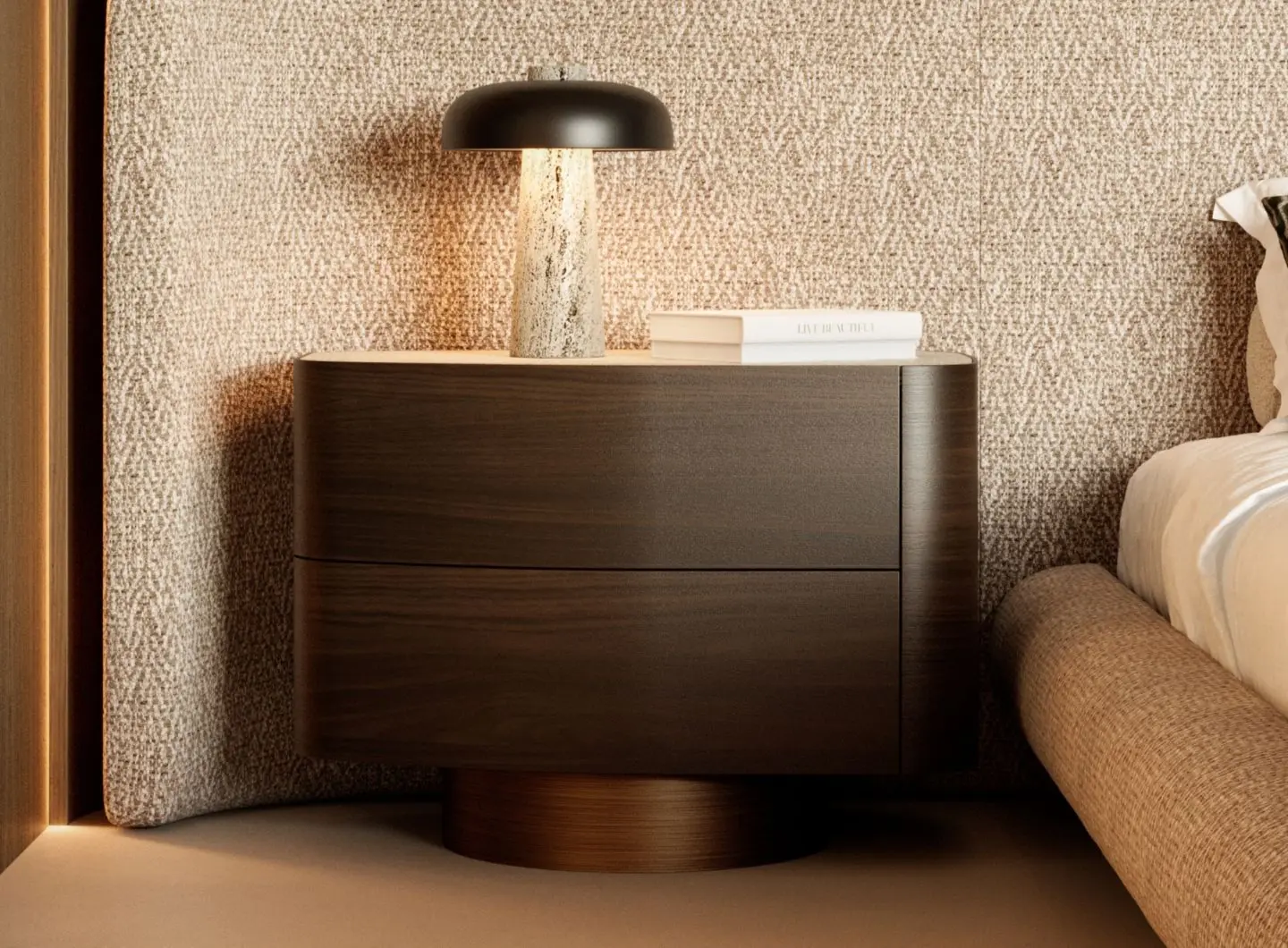 Il comodino Stone in legno scuro, dalla silhouette arrotondata e dotato di due cassetti, è abbinato a una lampada con finitura effetto pietra e a una pila di libri, e si trova accanto a un letto imbottito beige, davanti a una parete con un motivo discreto.
