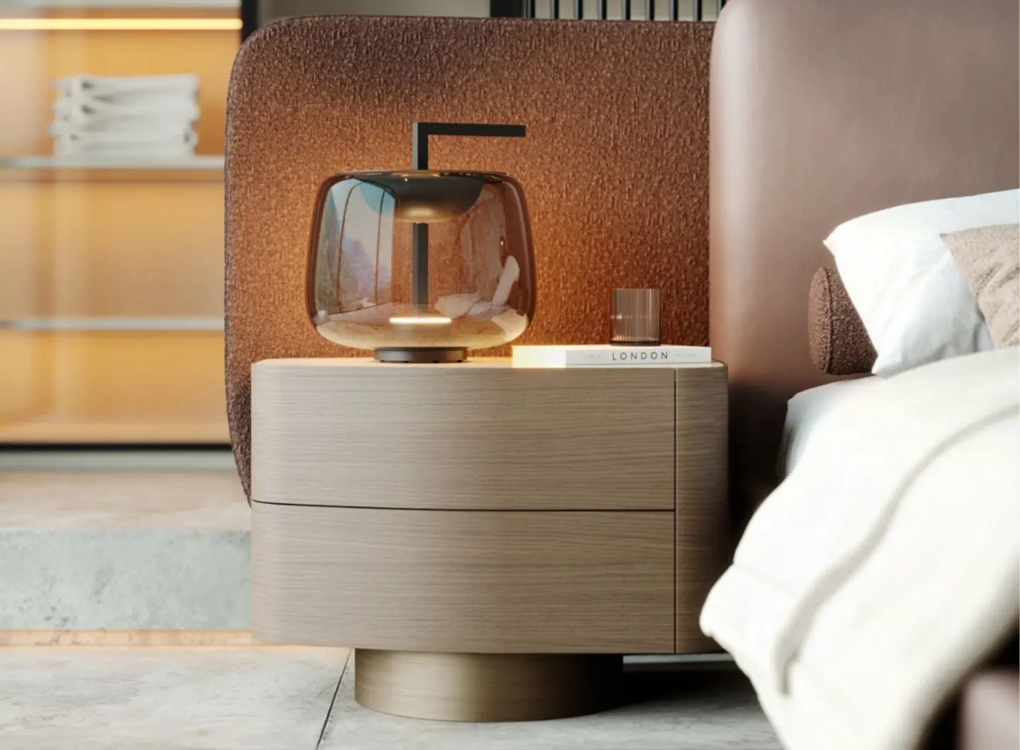 Il comodino Stone, in legno di noce chiaro con bordi arrotondati e due cassetti, è posizionato accanto a un letto con testiera imbottita marrone, impreziosito da una lampada di vetro, un bicchiere a coste e un libro in una camera da letto illuminata da luci soffuse.