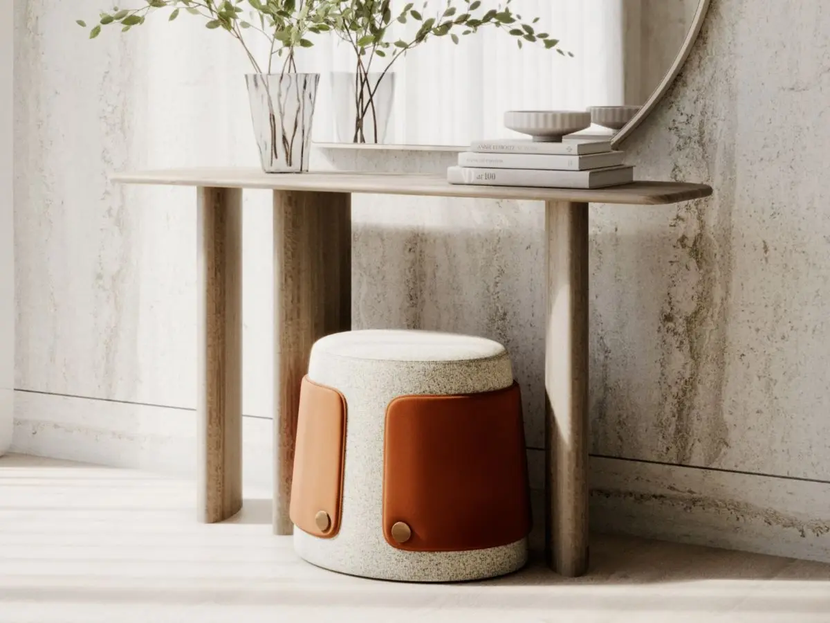 Lo Sgabello Dolomites appare sotto un tavolo consolle in legno come una seduta cilindrica dalla forma organica e arrotondata, rivestita in tessuto beige testurizzato con pannelli laterali in pelle marrone fissati da dettagli metallici rotondi, collocata in un ambiente minimalista dai materiali naturali.