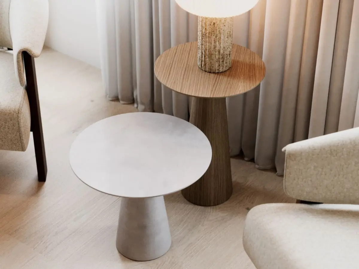 I tavolini Olivier appaiono affiancati in un ambiente minimalista, con il tavolino grigio in primo piano e quello in legno che sostiene una lampada materica, davanti a tende morbide e arredi neutri che esaltano le loro curve scultoree e l’eleganza nordica e discreta.
