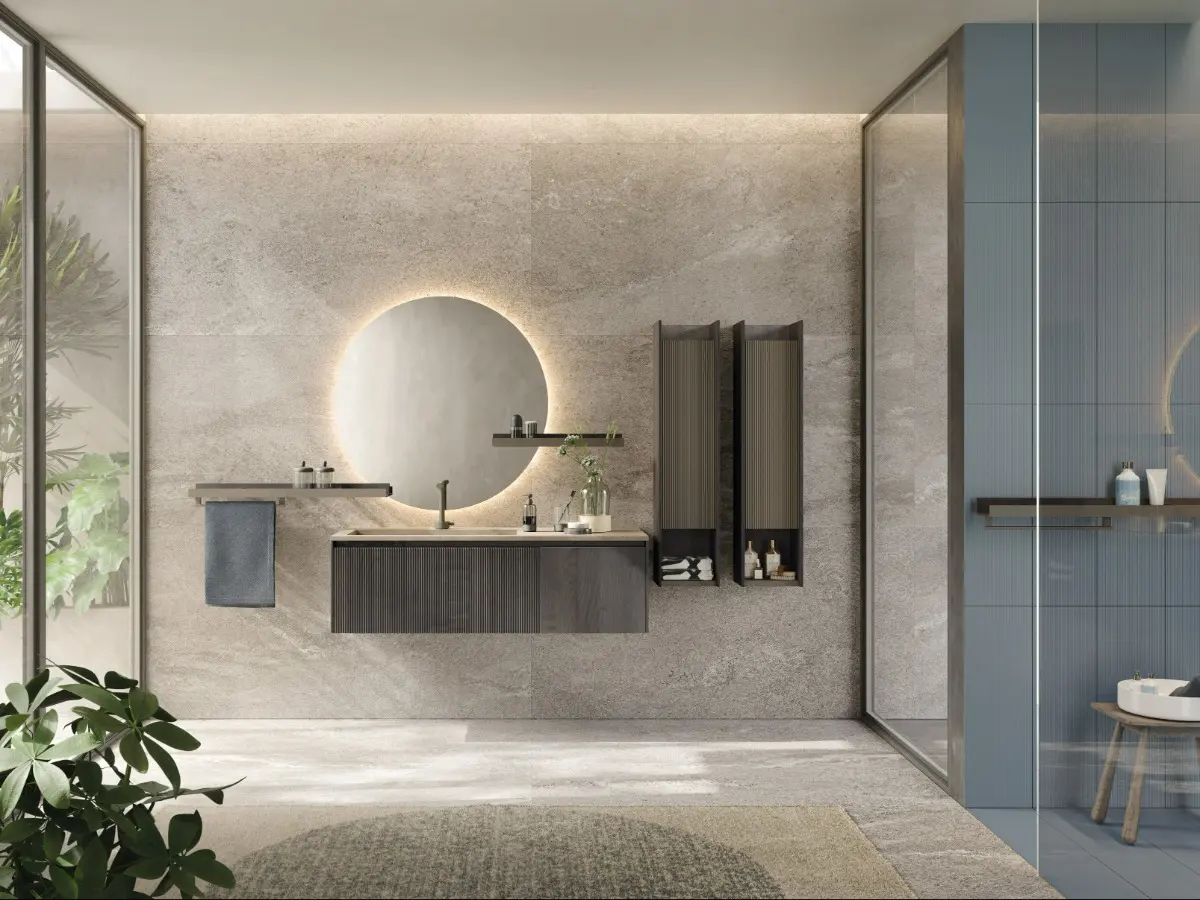 Bagno D'Art. Base portalavabo con anta plissé e anta liscia in rovere fumo, pensili con anta plissé in laccato lava opaco e struttura in rovere fumo, piano lavabo in gres porcellanato Fokos Roccia, mensole bicolor in laccato lava opaco e rovere fumo. Barra porta asciugamani, specchio tondo con led perimetrale.